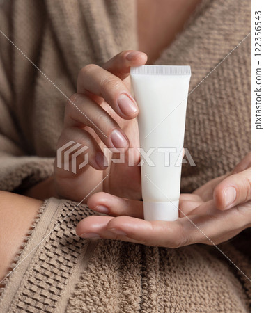 Closeup of woman in brown bathrobe holding squeeze cream tube in hands, cosmetic mockup 122356543