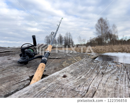 Fishing rod on a wooden dock with a serene water and natural landscape background. 122356858