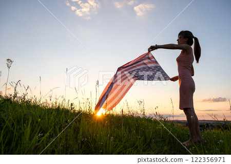 Back view of happy woman with USA national flag standing outdoors at sunset. Positive female celebrating United States independence day. International day of democracy concept. 122356971
