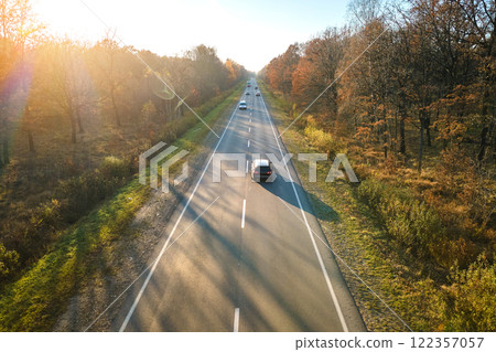 Aerial view of intercity road with fast driving cars between autumn forest trees at sunset. Top view from drone of highway traffic in evening Aerial view of intercity road with fast driving cars between autumn forest trees at sunset. Top view from drone of highway traffic in evening 122357057
