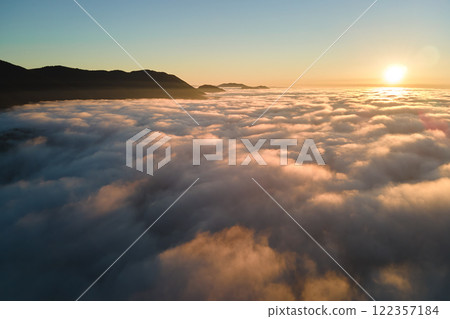 Aerial view of colorful sunrise over white dense fog with distant dark silhouettes of mountain hills on horizon 122357184