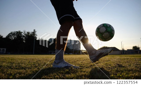 Legs of young man kicking ball at green field. Male feet of professional footballer juggling soccer ball on stadium at sunset. Sportsman practicing tricks outdoor. Concept of a freestyle football Legs of young man kicking ball at green field. Male feet of professional footballer juggling soccer ball on stadium at sunset. Sportsman practicing tricks outdoor. Concept of a freestyle football 122357554