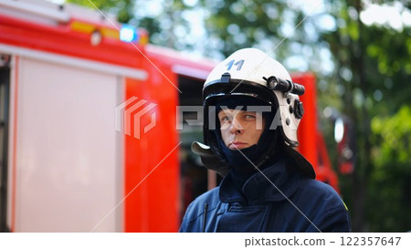 Portrait of confident male fireguard with dirty face standing near a fire engine. Young fireman in full equipment posing after fire against the background of big red truck. Concept of saving lives 122357647
