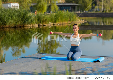 Sportive young woman exercising with dumbbells sitting on yoga mat near pond in park on sunny day Sportive young woman exercising with dumbbells sitting on yoga mat near pond in park on sunny day 122357959