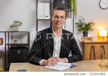 Young businessman looking to camera and writing down on diary in video chat at home office desk Young businessman looking to camera and writing down on diary in video chat at home office desk 122357985