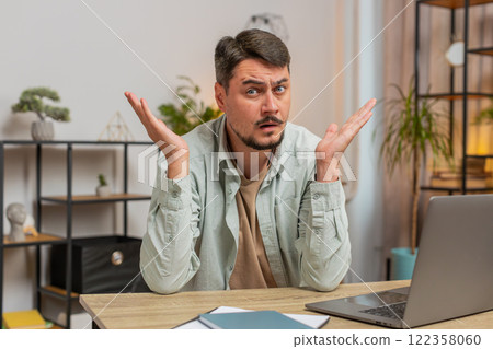 Angry young man raising hands in indignant expression asking reason of failure sitting at table Angry young man raising hands in indignant expression asking reason of failure sitting at table 122358060