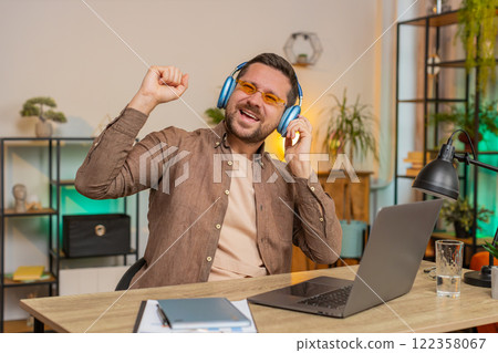Happy Caucasian man in headphones and sunglasses dancing listening to music at home office desk 122358067