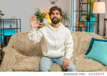 Happy young man vlogger smiling friendly at camera and waving hands while sitting on couch at home Happy young man vlogger smiling friendly at camera and waving hands while sitting on couch at home 122358175