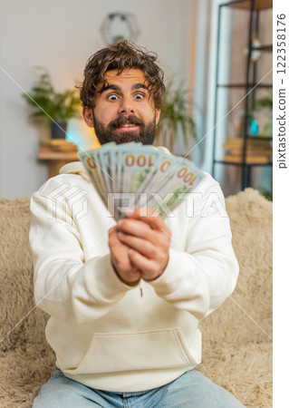 Successful rich young Hispanic man holding dollar bill money fan sitting on sofa couch at home Successful rich young Hispanic man holding dollar bill money fan sitting on sofa couch at home 122358176