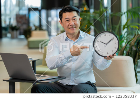 Mature businessman with laptop in lobby showing time on wall clock and pointing the finger at camera 122358337
