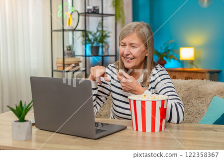 Mature woman watching movie on laptop with popcorn, laughing and enjoying content on sofa at home 122358367