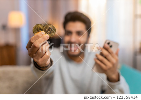 Young Indian man celebrating cryptocurrency win, showing bitcoin after checking exchange charts 122358414