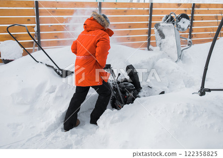 Red snow blower in action. Man outdoor in front of house using snowblower machine for removing snow on yard. Snow thrower in winter outside home. Young worker guy blowing snow 122358825