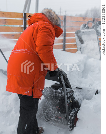 Red snow blower in action. Man outdoor in front of house using snowblower machine for removing snow on yard. Snow thrower in winter outside home. Young worker guy blowing snow 122358861