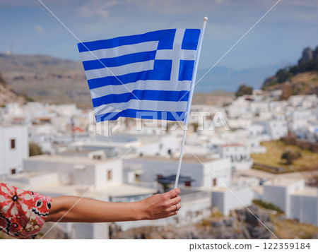 View of Greek flag waving in air serene Mediterranean vibe. beautiful Greek islands series, downtown Lindos, Greece 122359184