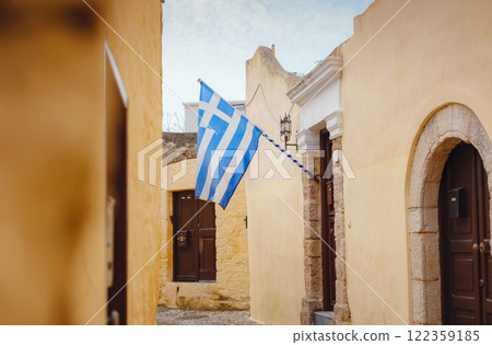 View of Greek flag waving in air serene Mediterranean vibe. beautiful Greek islands series, Narrow cobblestone street lined with charm in downtown Rhodes , Greece. Idyllic stone alley 122359185