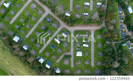 An aerial view of a campsite featuring neatly arranged pitches, caravans, motorhomes, and greenery. Pathways and surrounding trees add structure and nature to the scene. An aerial view of a campsite featuring neatly arranged pitches, caravans, motorhomes, and greenery. Pathways and surrounding trees add structure and nature to the scene. 122359483