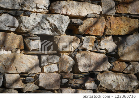 Sturdy stone wall showcasing a mix of textures in natural sunlight with shadows from nearby trees 122360736