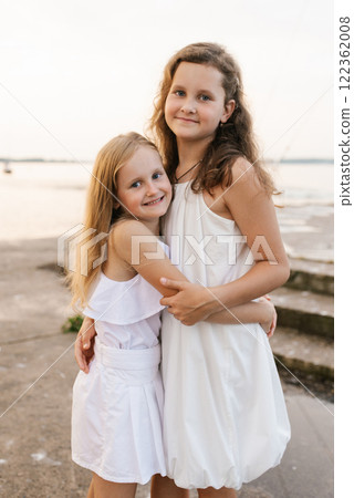 Two young girls are standing on beach, smiling and hugging each other 122362008