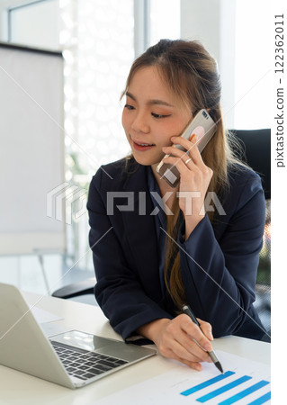 Businesswoman sitting at desk on couch in workplace or at home working on laptop and analyzing data on charts and graphs and writing on papers to make business plan and strategies for company. 122362011