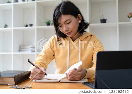 Intelligent young woman having online lessons on laptop, drinking coffee and taking notes, 122362485