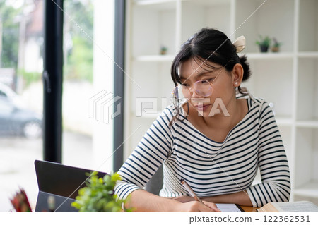 Intelligent young woman having online lessons on laptop, drinking coffee and taking notes, 122362531