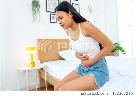 Side view of beautiful Asian woman sit on bed and express of stomach ache by use two hands to hold her stomach in bedroom. Side view of beautiful Asian woman sit on bed and express of stomach ache by use two hands to hold her stomach in bedroom. 122363280