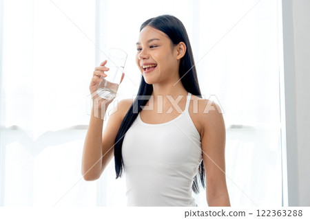 Close up beautiful Asian woman hold glass of water and stand in front of glass window with day light and she look happy with look at the glass. 122363288
