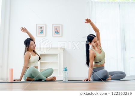 Pretty Asian women with exercise clothes do yoga posture with sit on yoga mat and raise their left arm over and cross head in room with day light. 122363289
