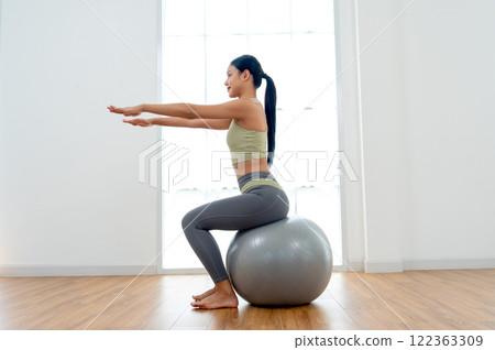 Pretty Asian woman with exercise clothes sit on exercise grey ball and raise arms forward and look her right direction with day light in front of glass window. 122363309