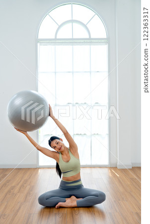 Vertical image of pretty Asian woman with exercise clothes sit and do yoga exercise posture by hold grey ball and raise to her right side and stay in front of glass window. 122363317