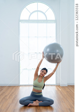 Vertical image of pretty Asian woman with exercise clothes sit and do yoga exercise posture by hold grey ball and raise to her left side and stay in front of glass window. Vertical image of pretty Asian woman with exercise clothes sit and do yoga exercise posture by hold grey ball and raise to her left side and stay in front of glass window. 122363318