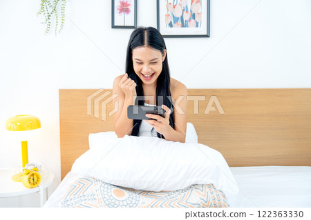Front view of beautiful Asian woman sit on bed and express success feeling and happiness during play game on her mobile phone with day light Front view of beautiful Asian woman sit on bed and express success feeling and happiness during play game on her mobile phone with day light 122363330