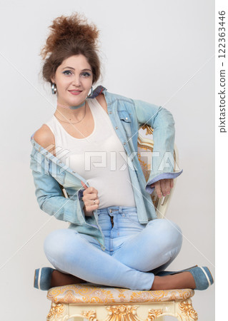 A beautiful woman in a denim suit poses on a chair on a gray background. 122363446