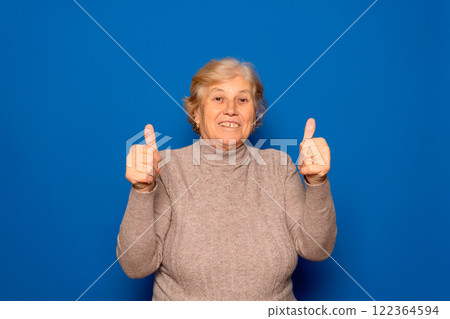 Caucasian mature woman about 70 years old wearing dark beige sweater with thumbs up in sign of positivity and satisfaction, isolated on blue studio background. 122364594