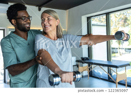 African American doctor helping patient treating arms with dumbbells during rehabilitation session 122365097