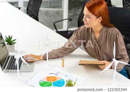 Young female engineer sitting at the table and looking at the wind turbine model Young female engineer sitting at the table and looking at the wind turbine model 122365394