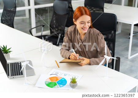 Young female engineer sitting at the table and looking at the wind turbine model Young female engineer sitting at the table and looking at the wind turbine model 122365397