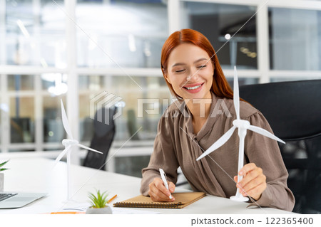 Young female engineer sitting at the table and looking at the wind turbine model Young female engineer sitting at the table and looking at the wind turbine model 122365400