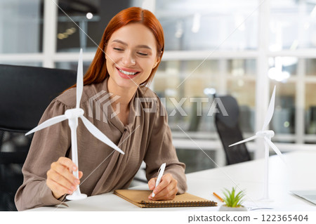 Young female engineer sitting at the table and looking at the wind turbine model 122365404