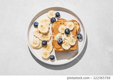 Peanut butter toast on a plate with banana and blueberry on a light background with shadow. Peanut butter toast on a plate with banana and blueberry on a light background with shadow. 122365789