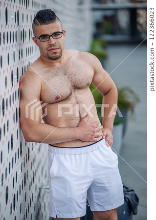 Strong sportsman with glasses and white shorts leaning against a wall and touching his abdomen 122366023