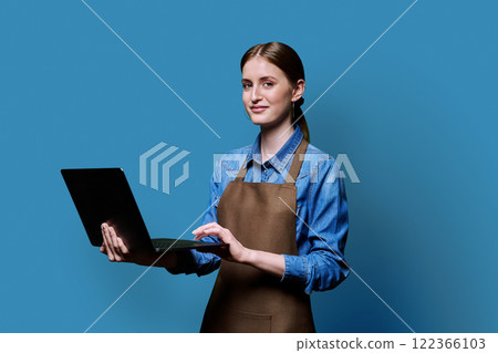Portrait of young woman in apron holding laptop on blue background 122366103