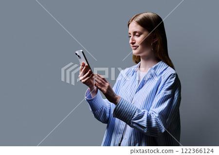 Teenage student girl in blue shirt holding smartphone in hands on grey studio background 122366124
