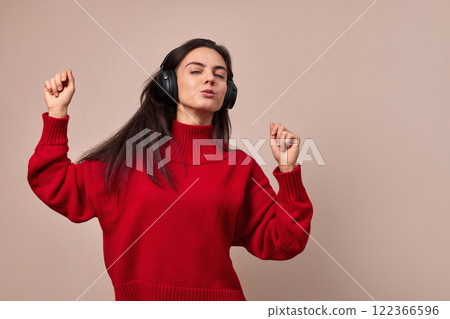 pretty girl wearing red sweater and headphones enjoying listening music and dancing on pink background. pretty girl wearing red sweater and headphones enjoying listening music and dancing on pink background. 122366596