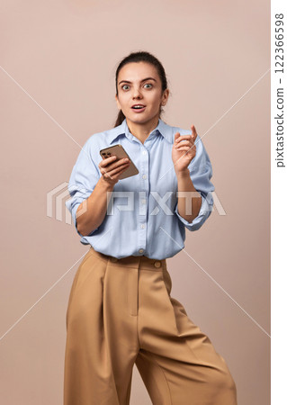 shocked amazed pretty woman holding mobile phone looking at camera on beige background. 122366598