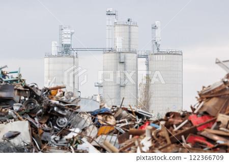 Massive concrete silos tower behind piles of scattered scrap metal and industrial debris. Scene shows impact of heavy manufacturing on fragile environment, urging sustainable waste solutions 122366709