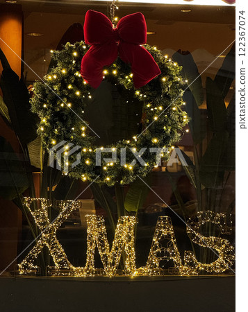 A beautifully adorned Christmas wreath with twinkling lights hangs prominently in a window display. Below it, illuminated letters spell out the word XMAS, enhancing the festive atmosphere. 122367074