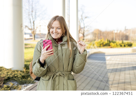 Smiling woman using mobile phone app, playing game, shopping online outdoors. Happy woman celebrating victory in lottery. Amazed hipster wondered with win in online auction betting on website Smiling woman using mobile phone app, playing game, shopping online outdoors. Happy woman celebrating victory in lottery. Amazed hipster wondered with win in online auction betting on website 122367451