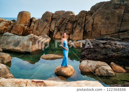 Stunning model in wet blue dress stands on rock at exotic ocean beach. Luxury fashion photoshoot. Natural water pool and rocky landscape. Perfect for high-end beauty, fragrance, travel ads. Stunning model in wet blue dress stands on rock at exotic ocean beach. Luxury fashion photoshoot. Natural water pool and rocky landscape. Perfect for high-end beauty, fragrance, travel ads. 122368886